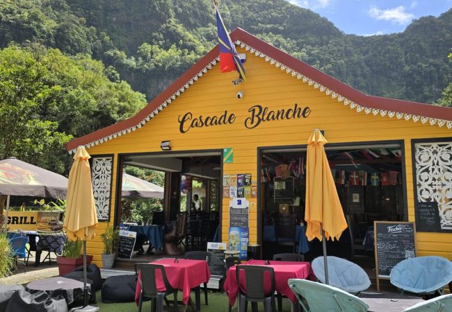 Жълтият дървен ресторант „Cascade Blanche“ с външни маси и чадъри, разположен пред стръмен планински склон.